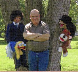 Photo of Daniel Jay in front of tree surrounded by his puppets